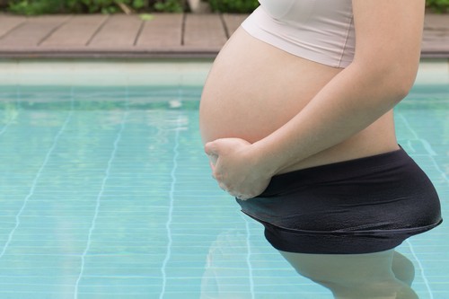 Pregnant woman hands holding belly on swimming pool background, healthy concept.